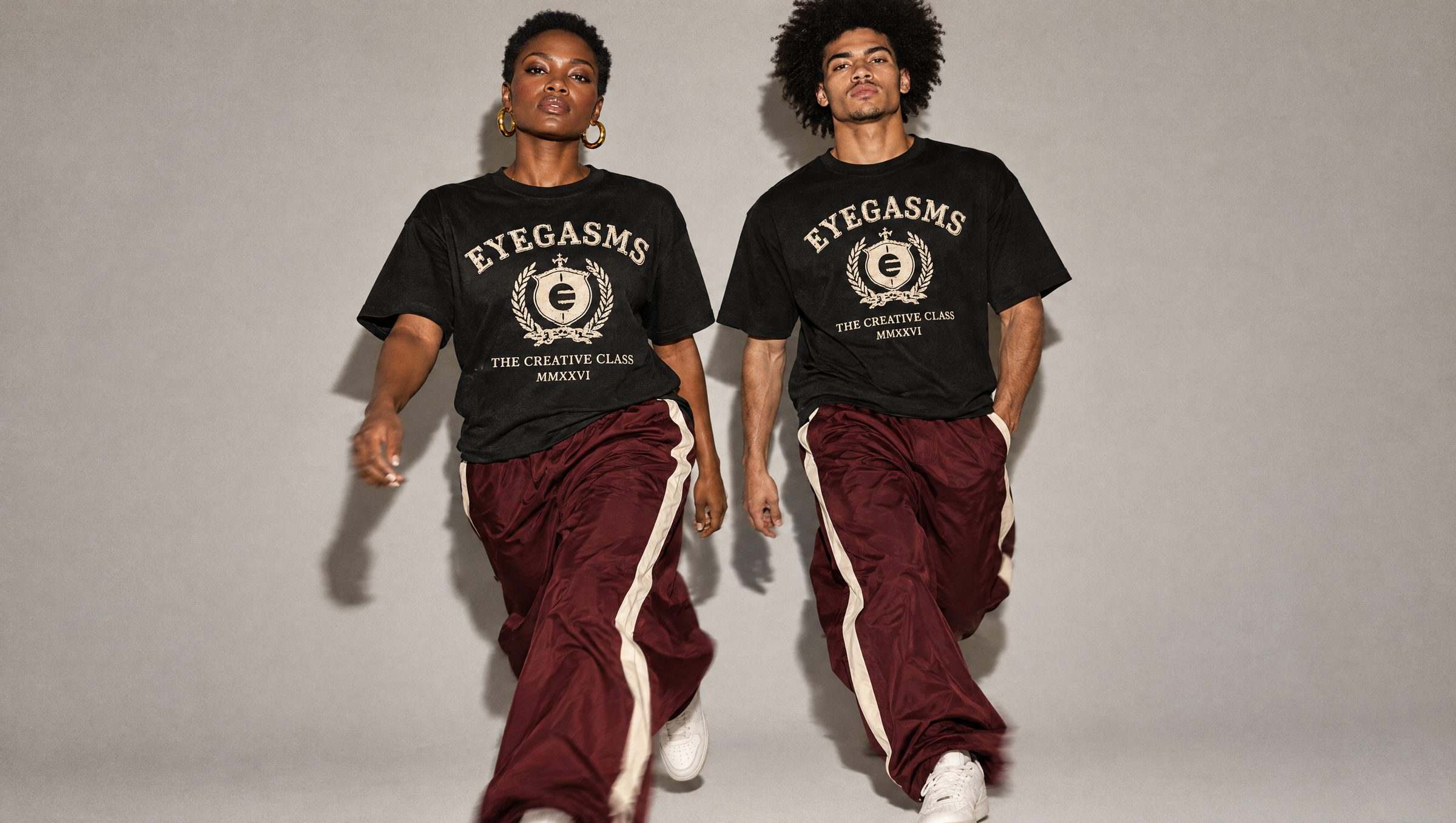 Two adults stride toward the camera against a flat grey backdrop in black EYEGASMS Creative Class tees, oxblood track trousers with a cream stripe and white sneakers, lit by direct flash.