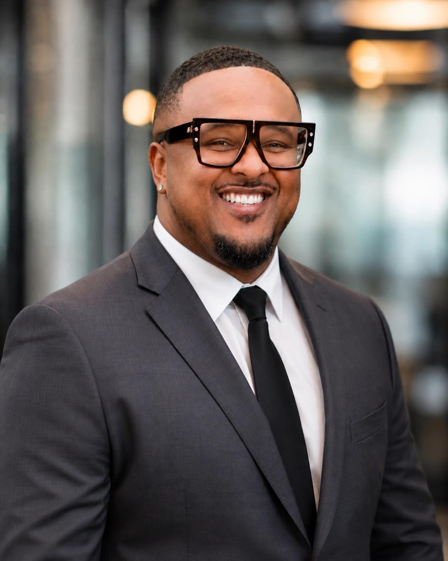Brennon Jackson, founder of EYEGASMS, smiling in a charcoal suit and bold tortoiseshell glasses.