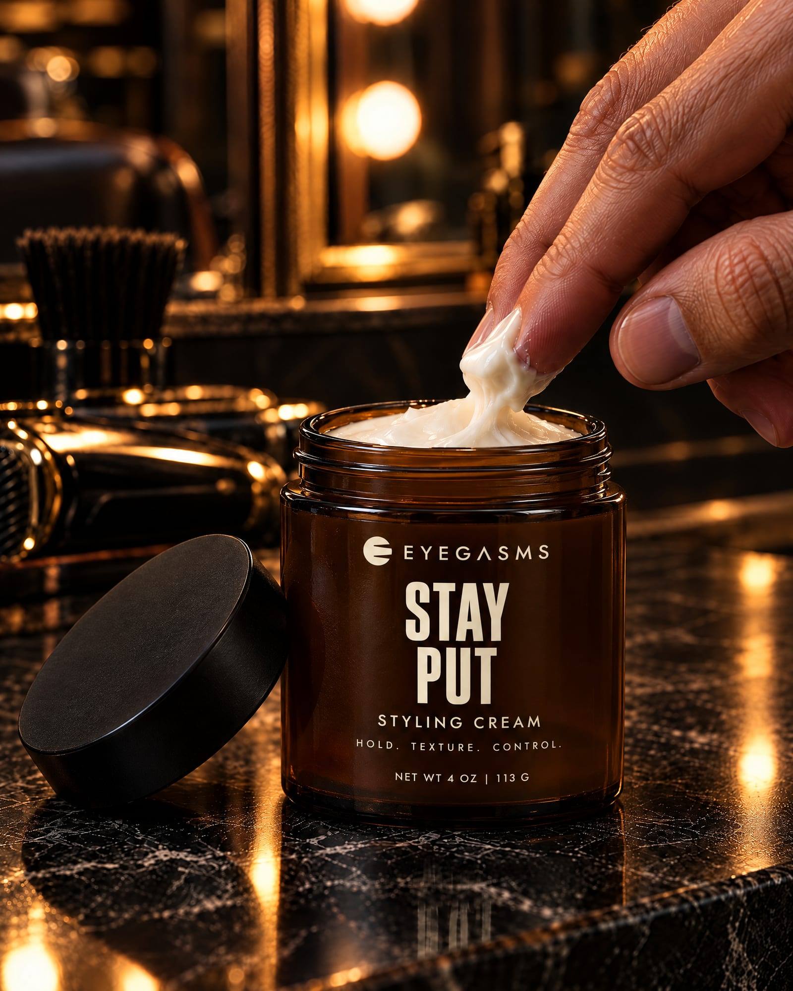 Stay Put, styling cream, by EYEGASMS: an open 4 oz amber glass jar of cream on a black marble barber counter, its black lid beside it, fingertips scooping a little out.