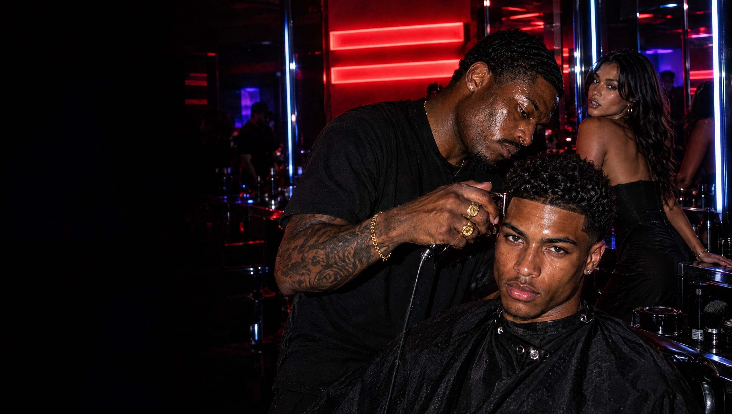 A tattooed barber with gold rings sharpens the line-up of a young man in a black cape who looks straight at the camera, while a woman in a black dress glances back from a mirror station; red neon bars and blue light glow behind them.