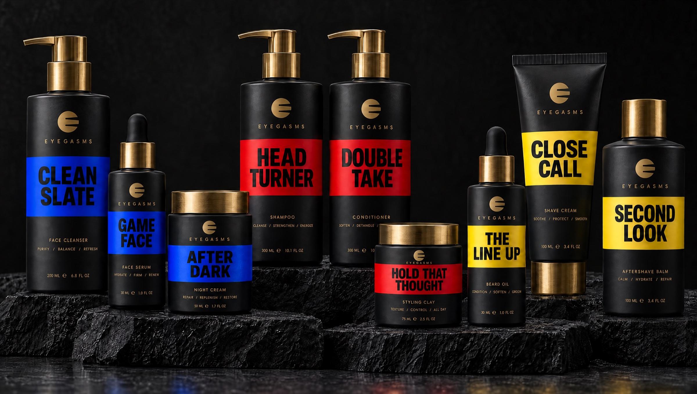 The full EYEGASMS range on black stone: nine matte black products with brass caps and bold color bands, cobalt for skincare (Clean Slate, Game Face, After Dark), red for hair care (Head Turner, Double Take, Hold That Thought) and yellow for grooming (The Line Up, Close Call, Second Look).