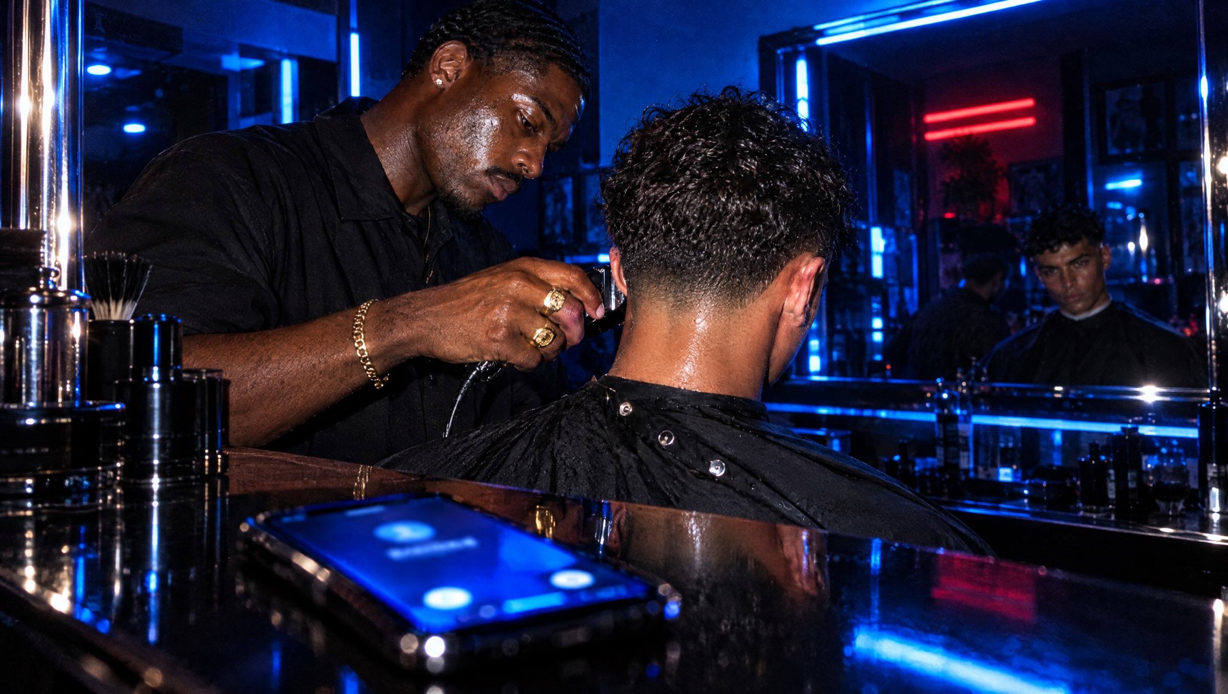 A phone lights up blue on a black lacquered station while, behind it, a barber keeps blending a fade under blue light, a red neon reflected in the mirror.