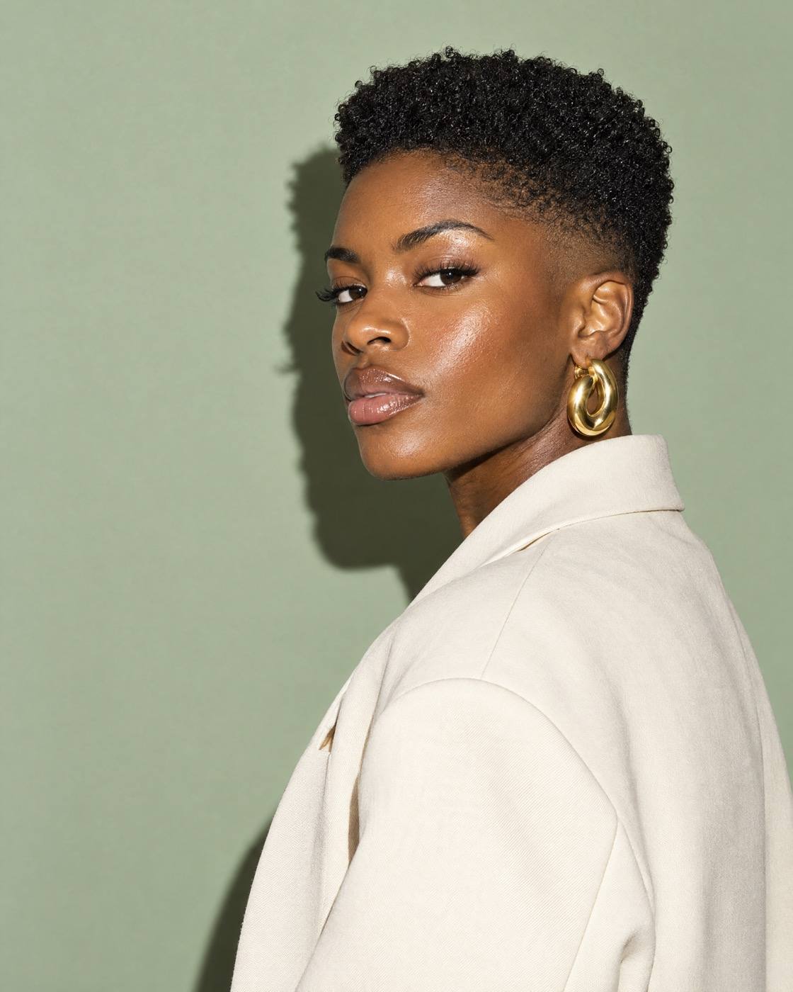 Studio portrait of a woman with a fresh tapered natural cut and gold earrings, in a cream blazer, on sage paper.