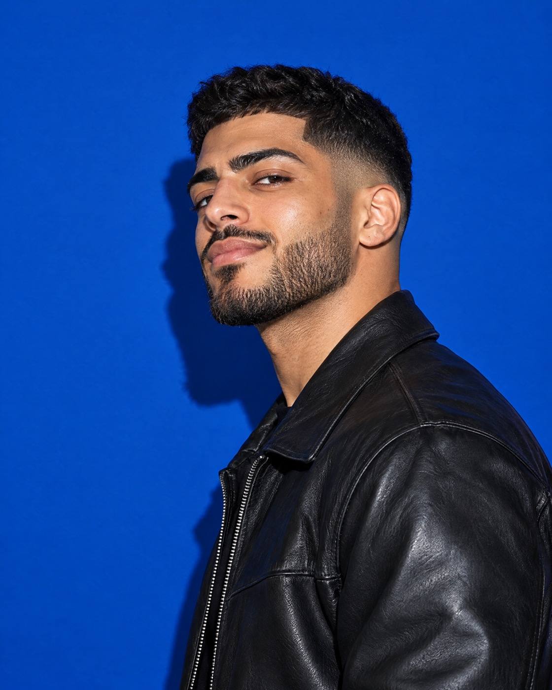 Studio portrait of a man with a sharp drop fade and trimmed beard in a leather jacket, on cobalt paper.