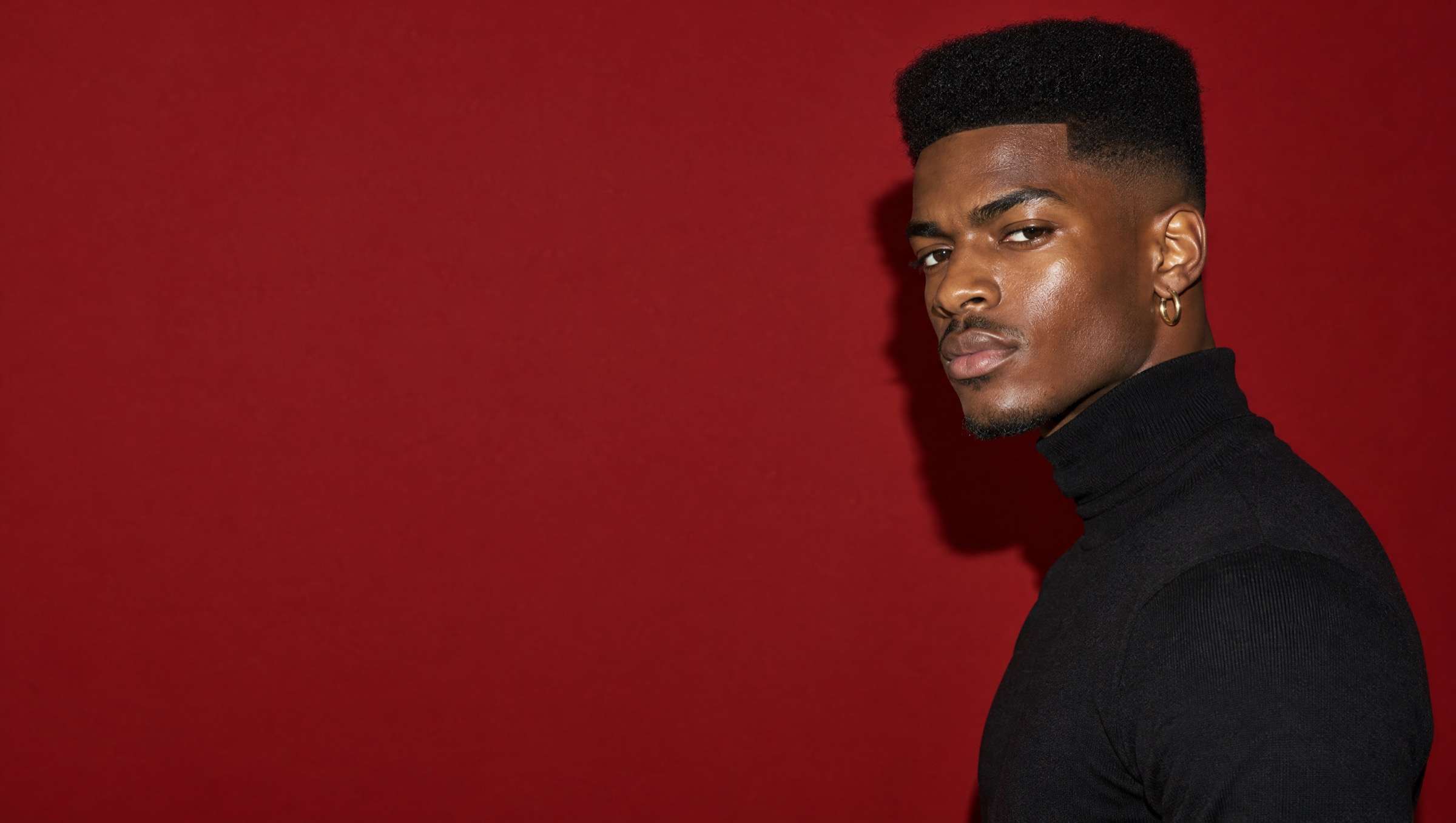 Studio portrait of a young man with a sharp high-top fade in a black turtleneck against deep oxblood paper.